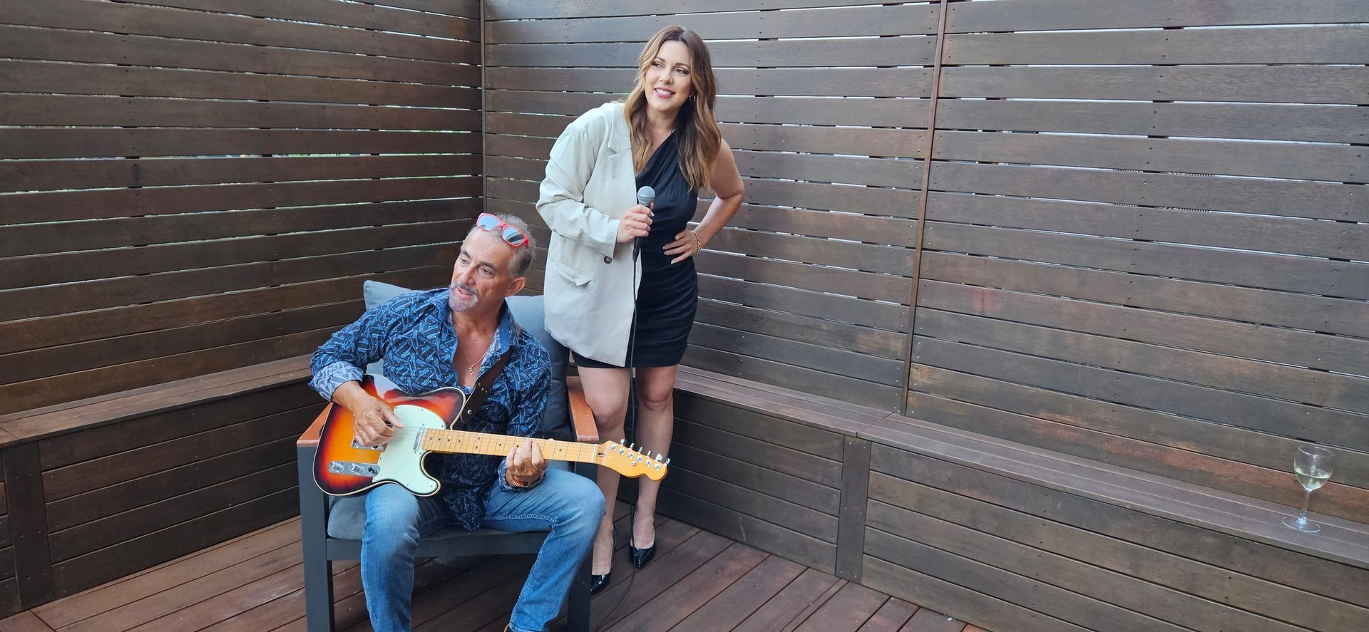 Person playing guitar sitting on a chair next to another person holding a microphone on a wooden deck.