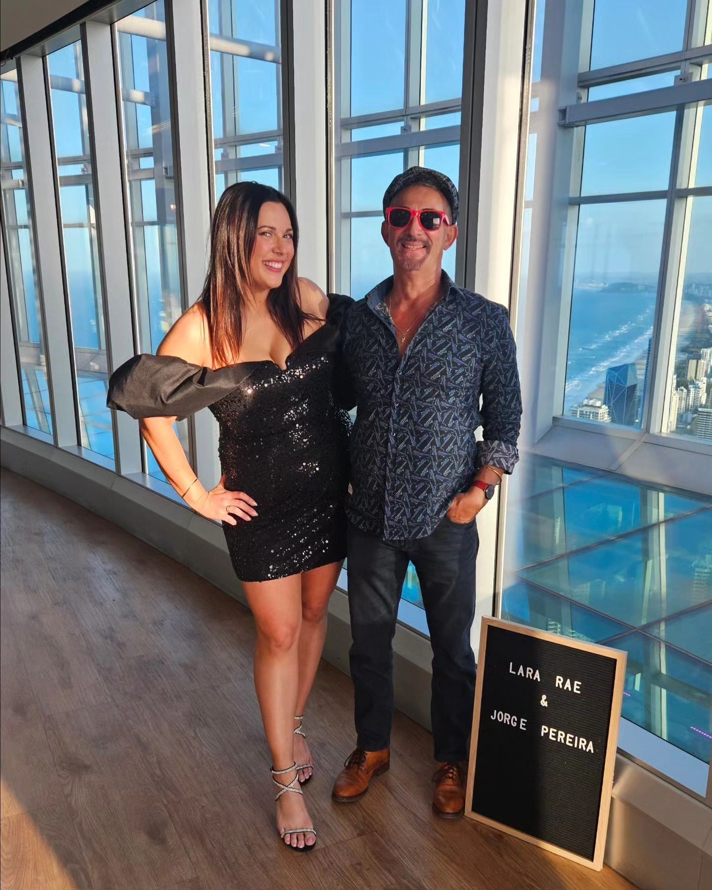 Man and woman posing in front of windows with city view, next to a sign with names.