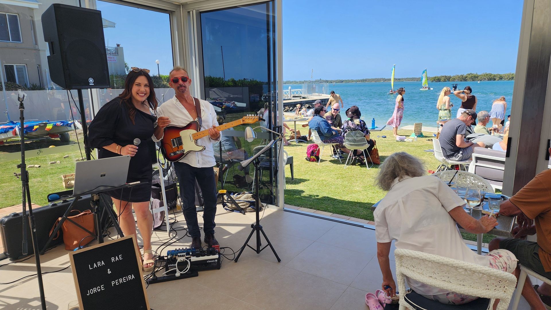 Musicians performing indoors with a view of people relaxing by the seaside under a clear blue sky.