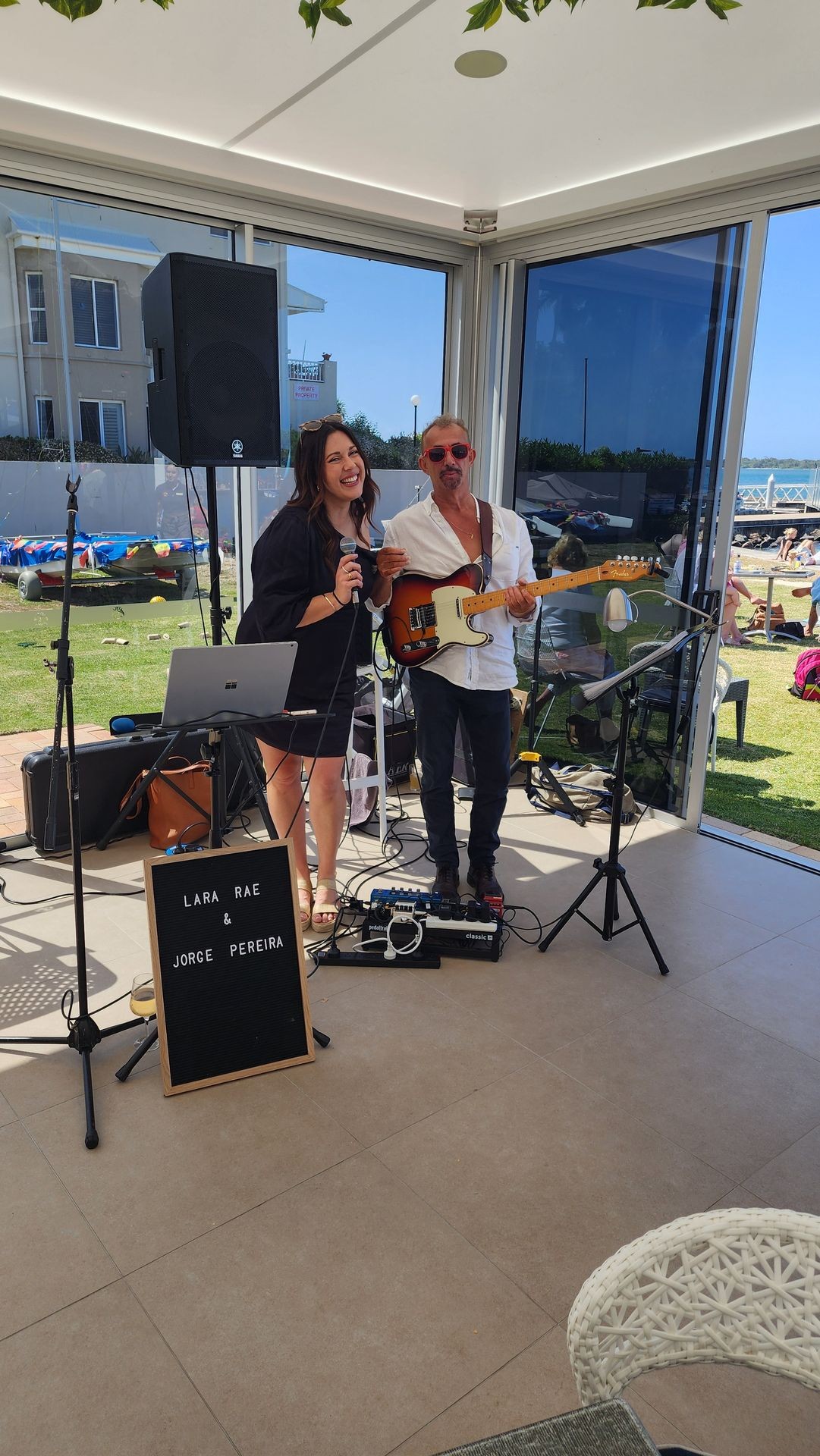 Two musicians performing with a guitar and microphone in a sunlit room by the seaside.